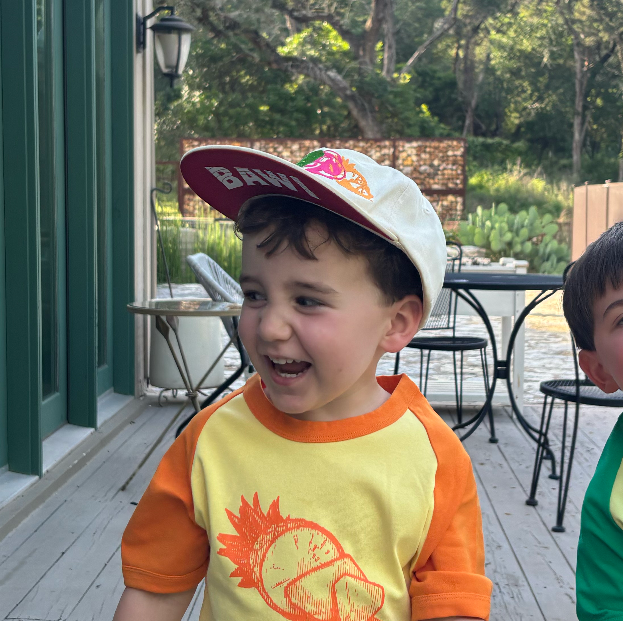 Child wearing a cap and colorful shirt on a wooden deck with outdoor furniture and greenery.